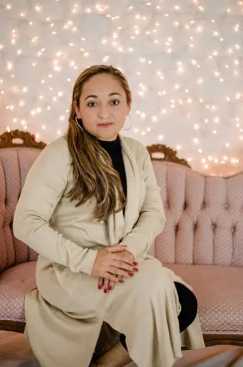 A person with long hair sitting on a pink tufted couch in front of a wall decorated with warm, glowi…