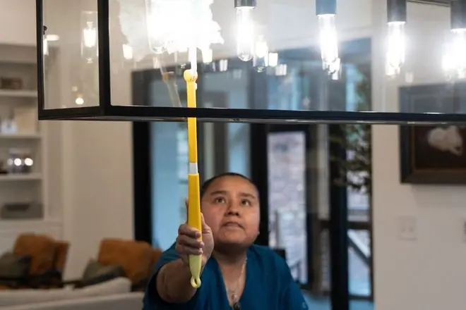 A person wearing a blue shirt uses a yellow duster to clean a rectangular glass light fixture hangin…