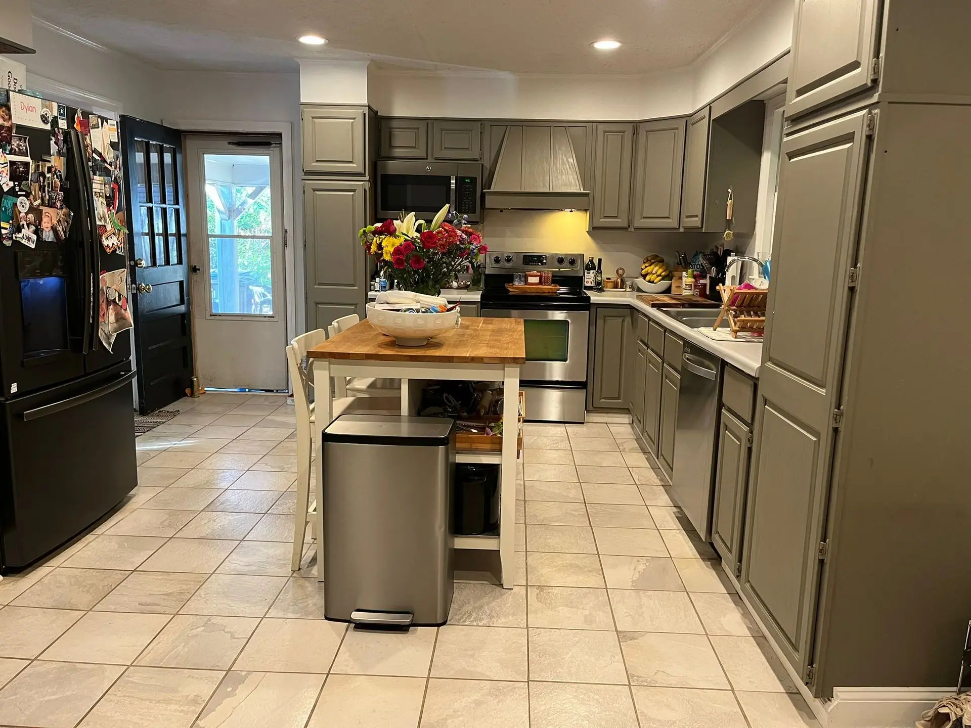 A kitchen with grey cabinets, a wooden island with white chairs, a stainless steel trash can, and light-colored tile floors.