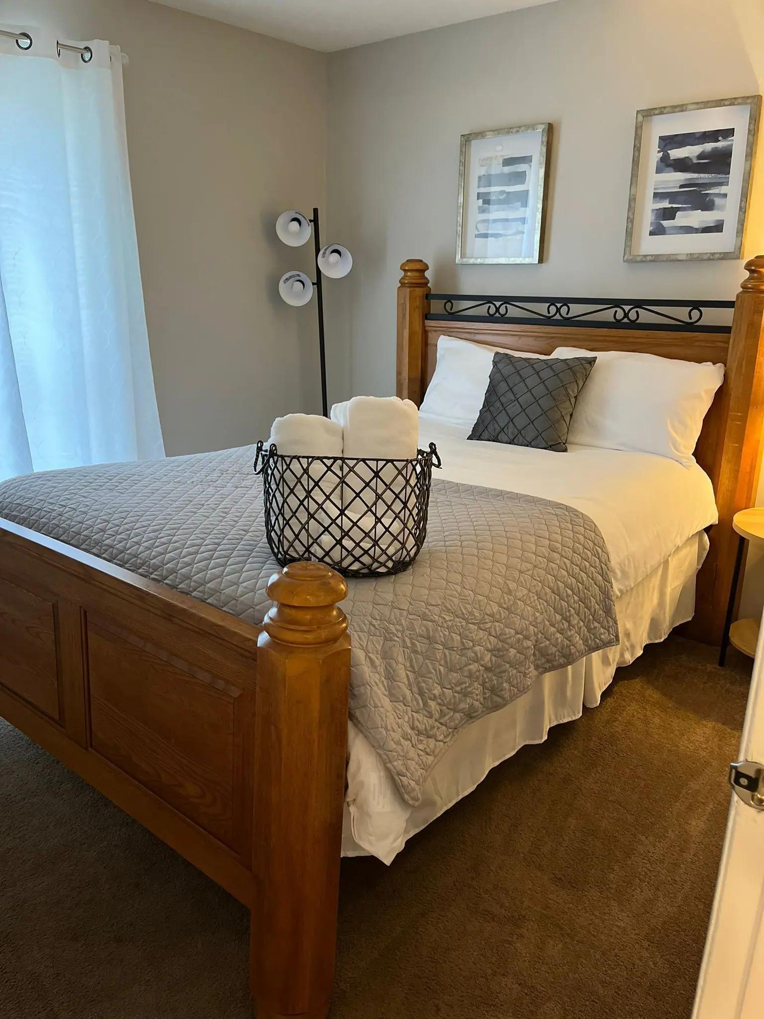 A wooden bed with a grey textured quilt and a black wire basket of towels, set in a neutral-toned bedroom with wall art.