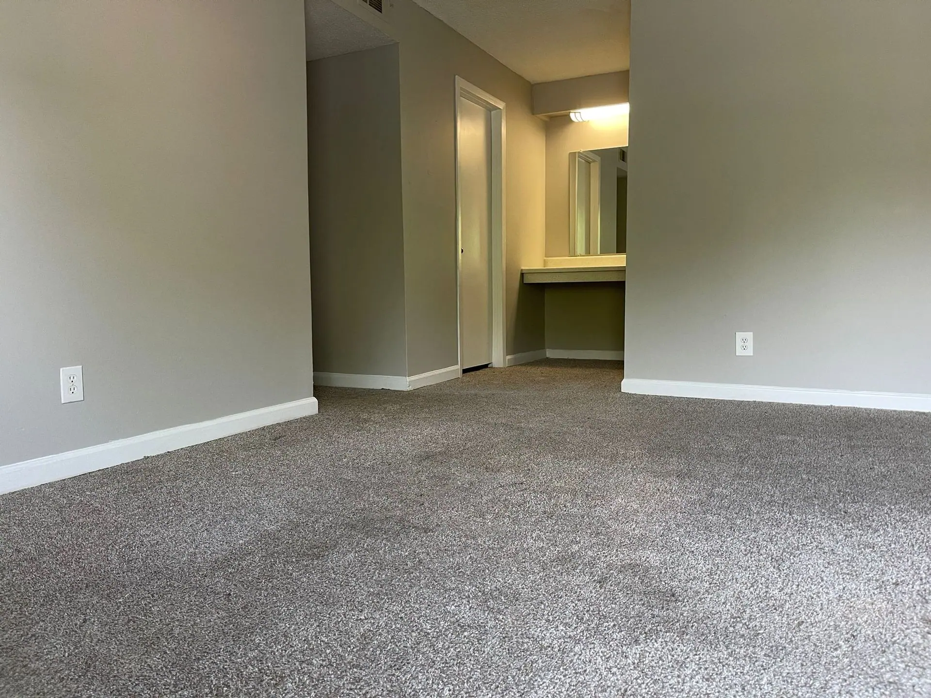 An interior room featuring light gray walls, gray speckled carpeting, a doorway to the left, and a vanity area ahead.
