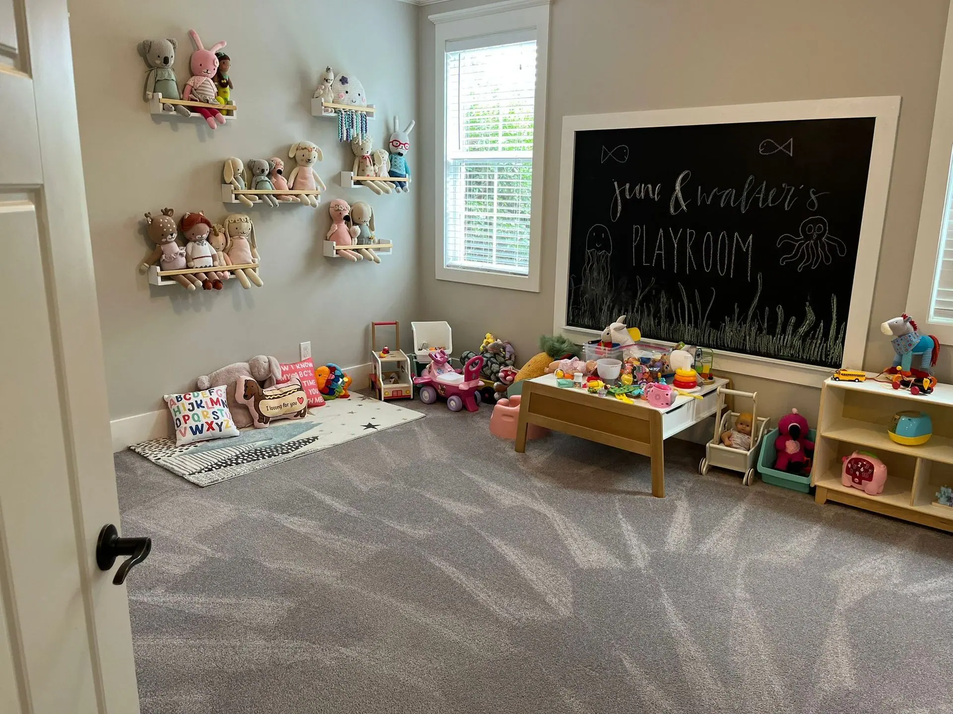 A cozy playroom with gray carpet, walls featuring plush toy displays, a large wall-mounted chalkboard, and toy furniture.