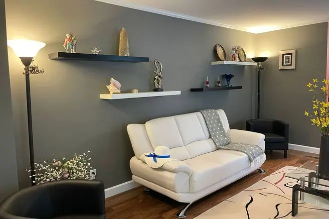 A grey-walled living room features a white sofa, dark armchairs, floating wall shelves with decor, a…